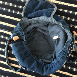 Puede incluir: Una bolsa de cord&oacute;n de mezclilla azul con una solapa superior y una sola correa. La bolsa est&aacute; abierta y el interior es visible.