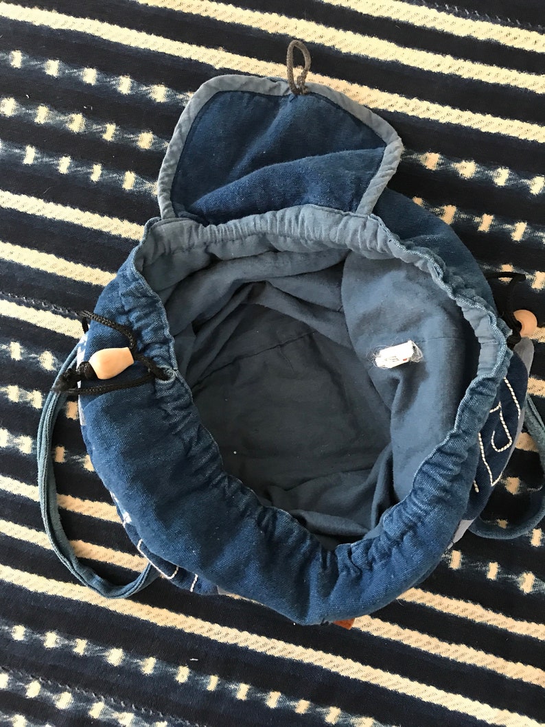 Puede incluir: Una bolsa de cord&oacute;n de mezclilla azul con una solapa superior y una sola correa. La bolsa est&aacute; abierta y el interior es visible.