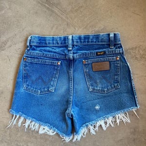 May include: Blue denim shorts with a frayed hem, featuring a high-rise waist, belt loops, and two back pockets. The right pocket displays a brown leather Wrangler brand patch, and both pockets feature the signature W stitching in dark thread.