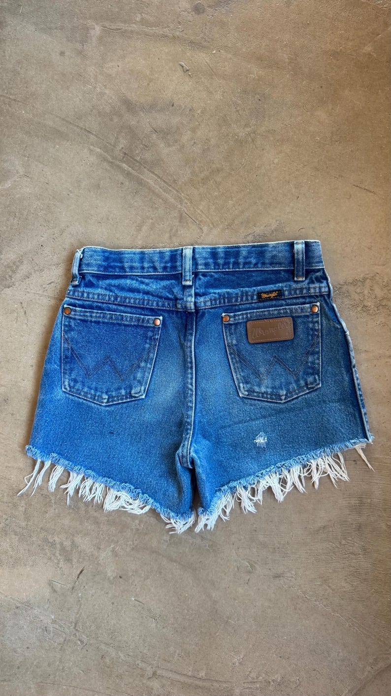 May include: Blue denim shorts with a frayed hem, featuring a high-rise waist, belt loops, and two back pockets. The right pocket displays a brown leather Wrangler brand patch, and both pockets feature the signature W stitching in dark thread.