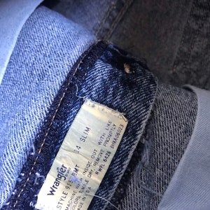 May include: A close-up of a blue denim Wrangler jeans tag with white text. The tag reads "Wrangler Style 9150 BMT 14 Slim Machine Wash Inside Out With Like Colors, Tumble Dry, Remove Promptly RWPL 6428 8HKA06023 Made in USA".