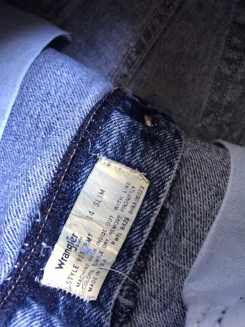 May include: A close-up of a blue denim Wrangler jeans tag with white text. The tag reads "Wrangler Style 9150 BMT 14 Slim Machine Wash Inside Out With Like Colors, Tumble Dry, Remove Promptly RWPL 6428 8HKA06023 Made in USA".