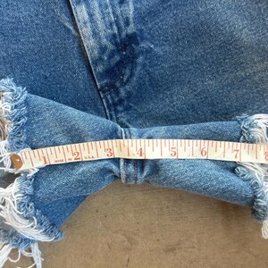 May include: Blue denim shorts featuring a frayed hemline and a classic five-pocket construction. A white measuring tape is positioned across the width of the leg opening, indicating a measurement of approximately 7.5 inches across the fabric.