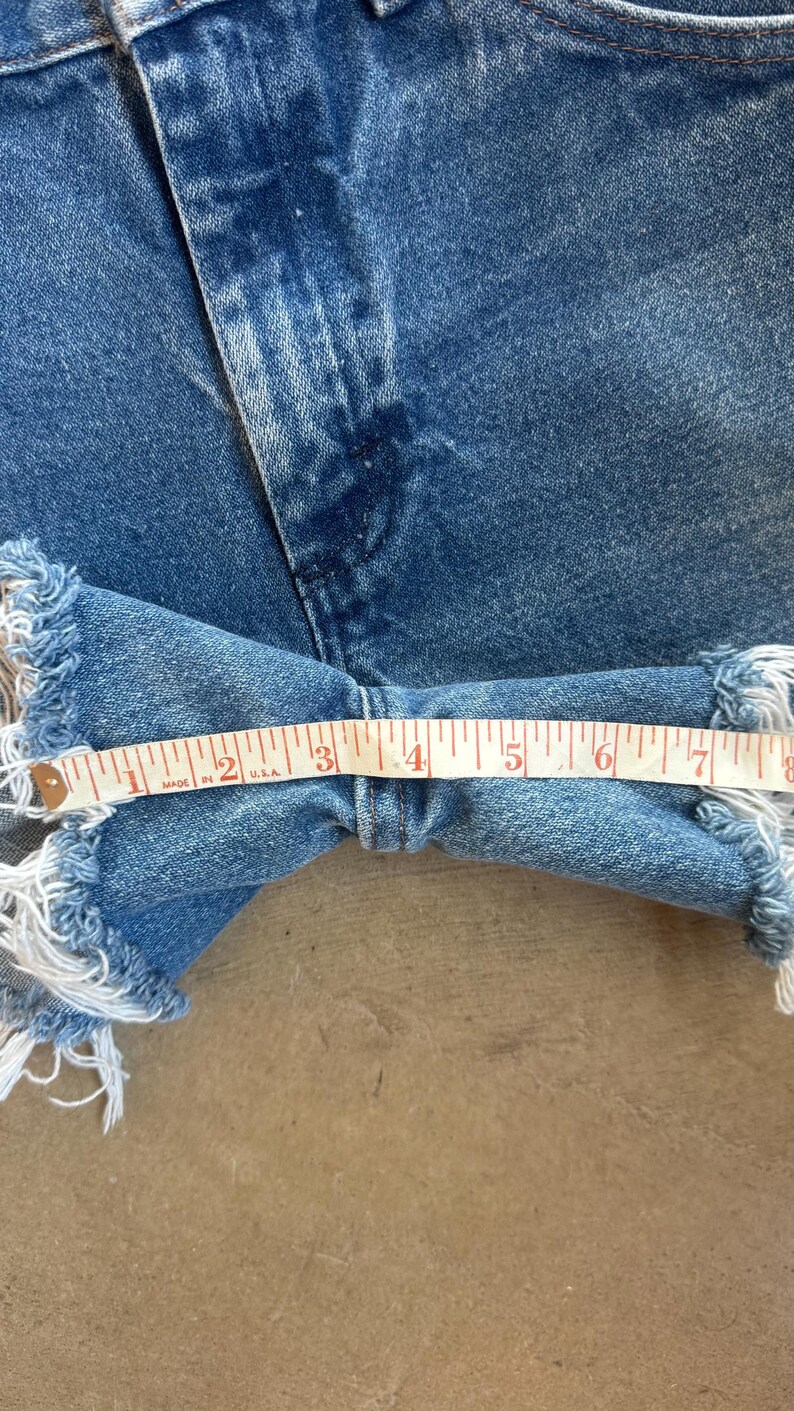 May include: Blue denim shorts featuring a frayed hemline and a classic five-pocket construction. A white measuring tape is positioned across the width of the leg opening, indicating a measurement of approximately 7.5 inches across the fabric.