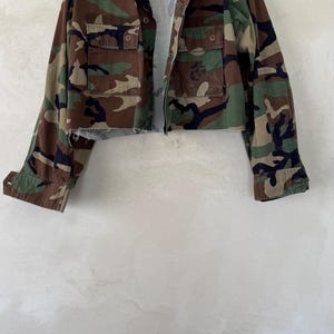 May include: A cropped jacket featuring a woodland camouflage pattern in shades of green, brown, and black. The jacket has a collared neckline, long sleeves with buttoned cuffs, and two front chest pockets with button closures. The hem is raw and frayed.
