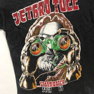 May include: Black t-shirt with a red and white graphic of a person wearing a gas mask and holding binoculars. The text "JETRO TULL...STORMWATCH...TOUR '80" is printed on the shirt.