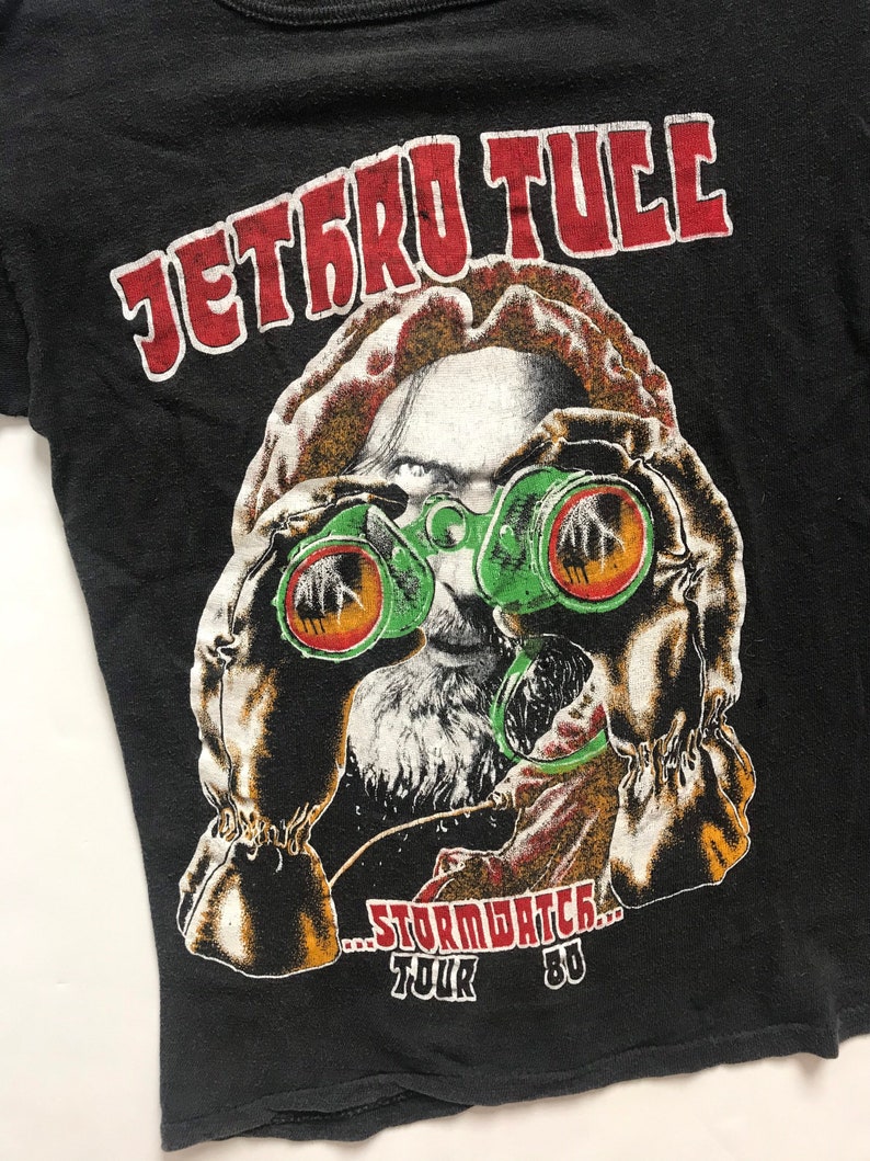 May include: Black t-shirt with a red and white graphic of a person wearing a gas mask and holding binoculars. The text "JETRO TULL...STORMWATCH...TOUR '80" is printed on the shirt.
