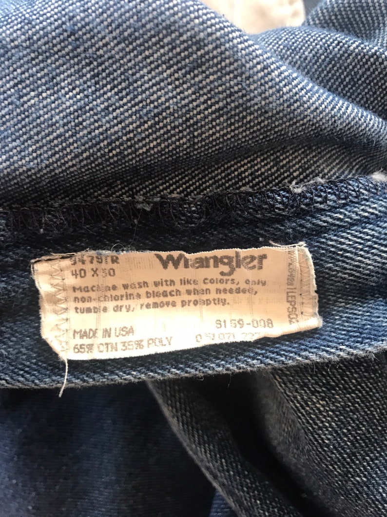 May include: A blue denim Wrangler jeans label with white text. The label says "9479TR 40 X 30 Wrangler Machine wash with like colors, only non-chlorine bleach when needed, tumble dry, remove promptly. MADE IN USA 3159-008 65% CTN 35% POLY".