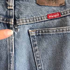 Pu&ograve; includere: Un paio di jeans blu denim con una toppa in pelle marrone che dice "Wrangler Authentic Jeans" e una toppa rossa che dice "Wrangler".