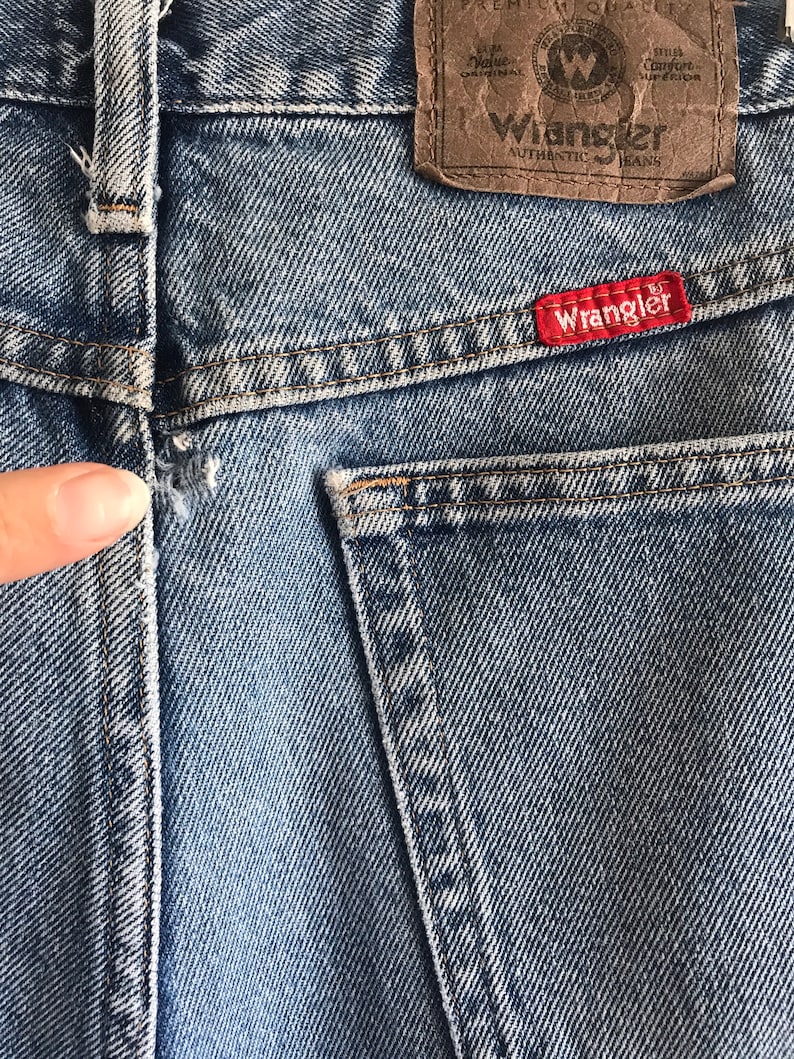 Pu&ograve; includere: Un paio di jeans blu denim con una toppa in pelle marrone che dice "Wrangler Authentic Jeans" e una toppa rossa che dice "Wrangler".
