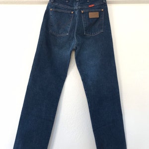 Może przedstawiać: Dżinsy z ciemnego denimu z brązową sk&oacute;rzaną naszywką na tylnej kieszeni z napisem "Wrangler".
