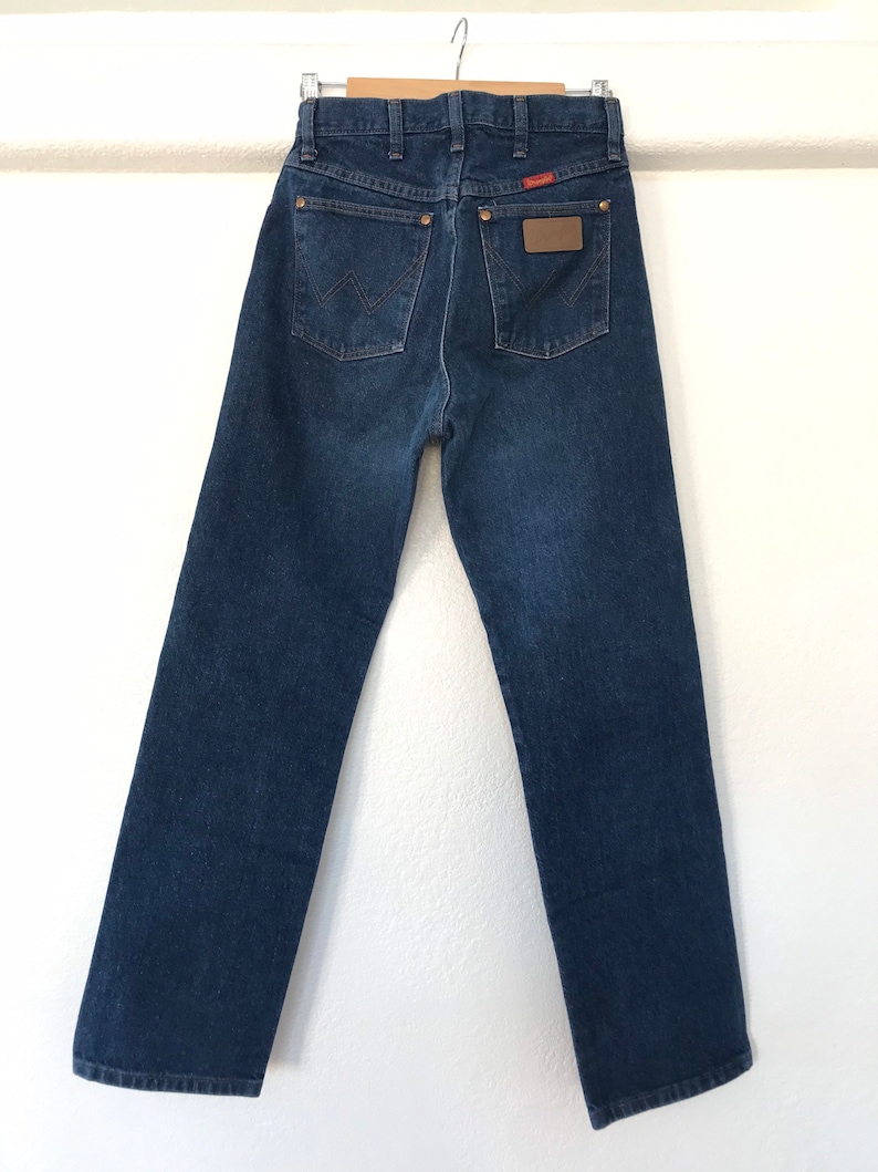 Może przedstawiać: Dżinsy z ciemnego denimu z brązową sk&oacute;rzaną naszywką na tylnej kieszeni z napisem "Wrangler".