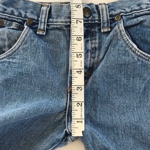 May include: A close-up of a pair of blue denim jeans with a measuring tape placed vertically along the front of the waistband. The tape measure reads from 1 to 10 inches.