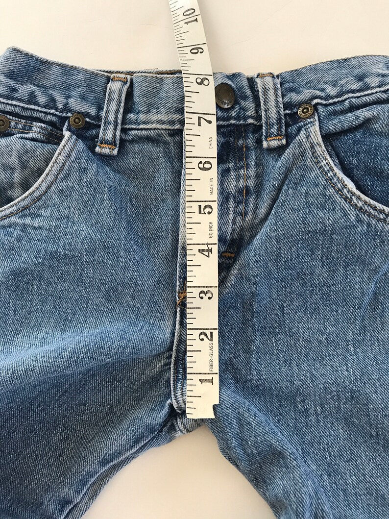 May include: A close-up of a pair of blue denim jeans with a measuring tape placed vertically along the front of the waistband. The tape measure reads from 1 to 10 inches.
