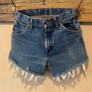 May include: Blue denim shorts featuring a classic five-pocket design, a button and zipper fly closure, copper-toned rivets, and a raw, frayed hemline with visible white threads hanging from the bottom edges of the legs.
