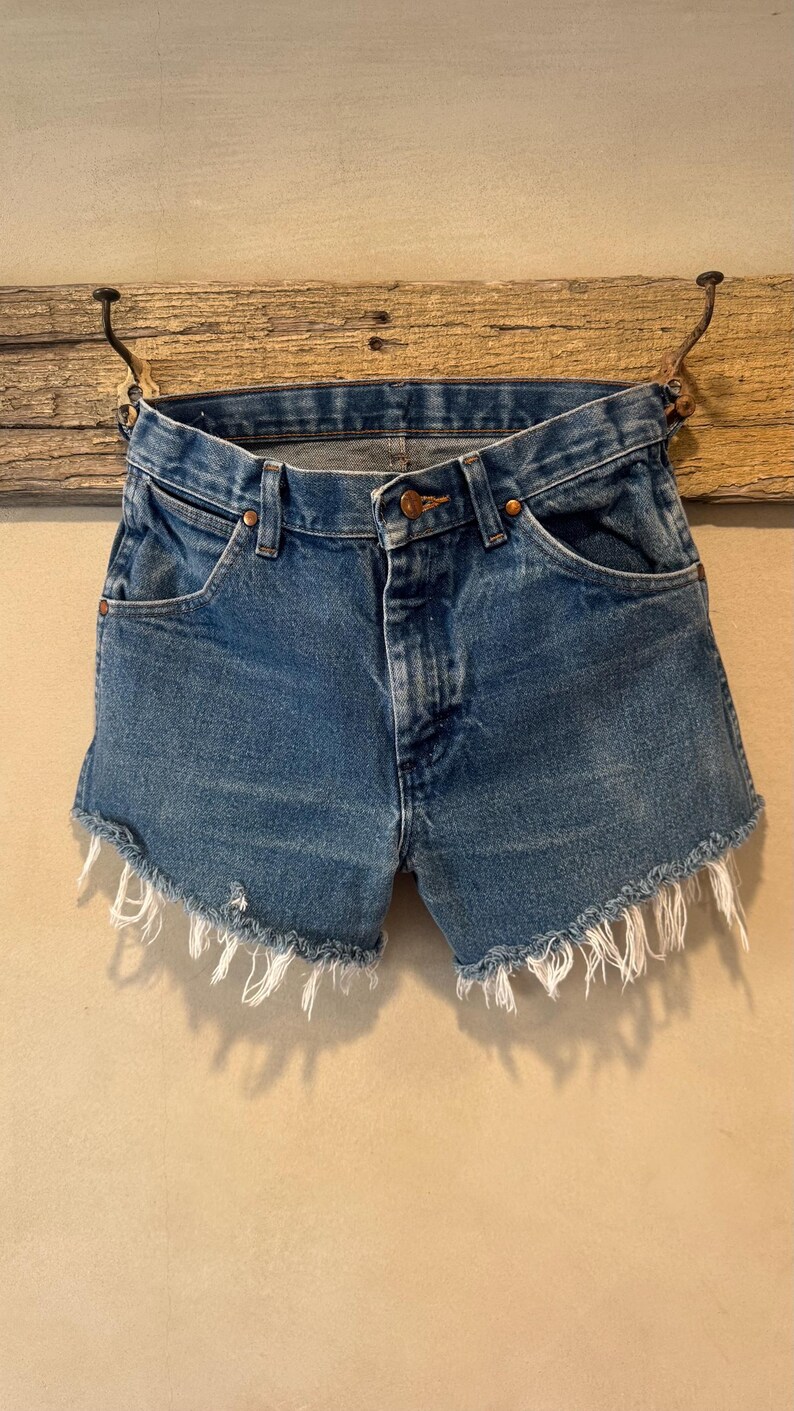 May include: Blue denim shorts featuring a classic five-pocket design, a button and zipper fly closure, copper-toned rivets, and a raw, frayed hemline with visible white threads hanging from the bottom edges of the legs.