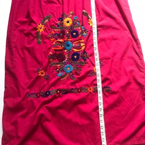May include: A long, pink dress with colourful floral embroidery. The dress has a gathered neckline and a loose, flowing fit. The embroidery is detailed and intricate, featuring a variety of flowers and leaves in shades of red, orange, yellow, green, blue, and purple.