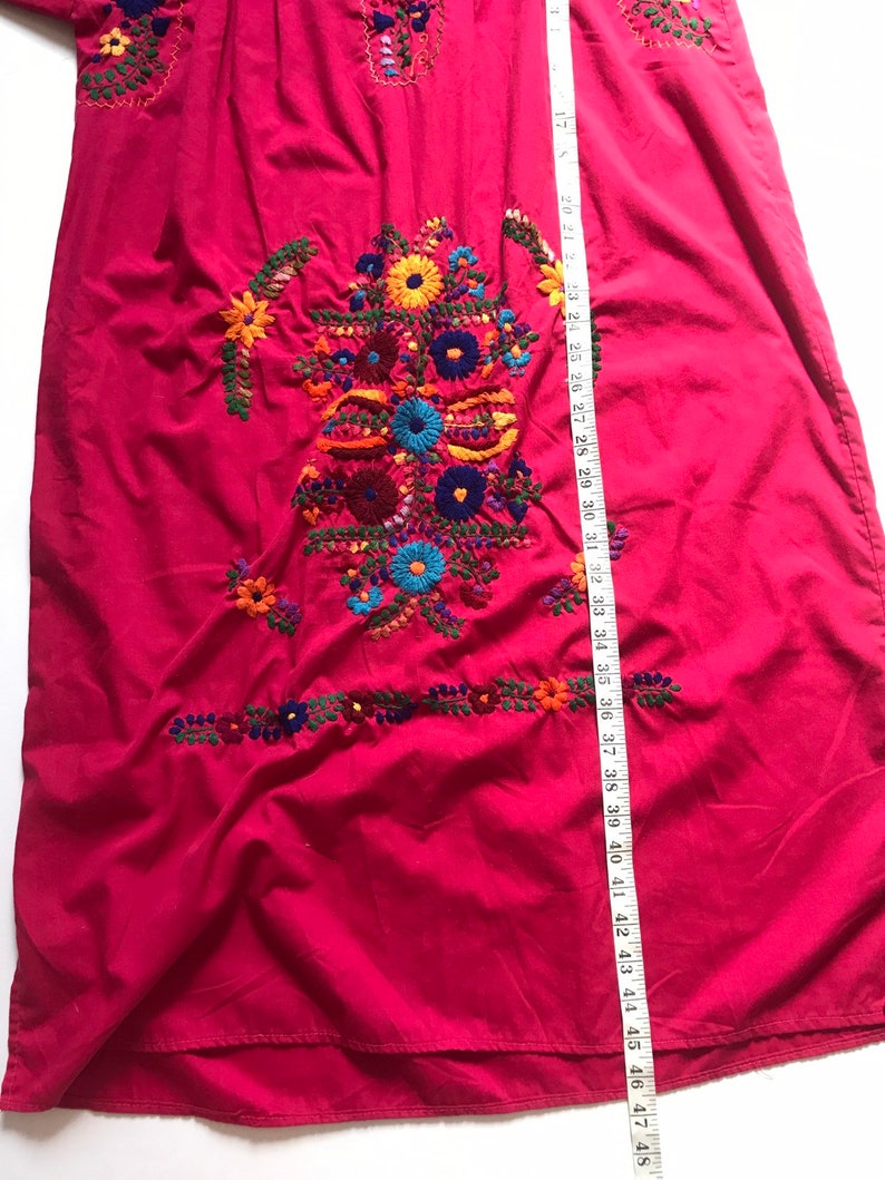 May include: A long, pink dress with colourful floral embroidery. The dress has a gathered neckline and a loose, flowing fit. The embroidery is detailed and intricate, featuring a variety of flowers and leaves in shades of red, orange, yellow, green, blue, and purple.