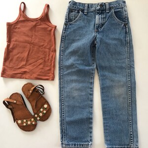 May include: A pair of brown leather sandals with white floral designs, a rust-colored tank top, and a pair of light blue denim jeans.