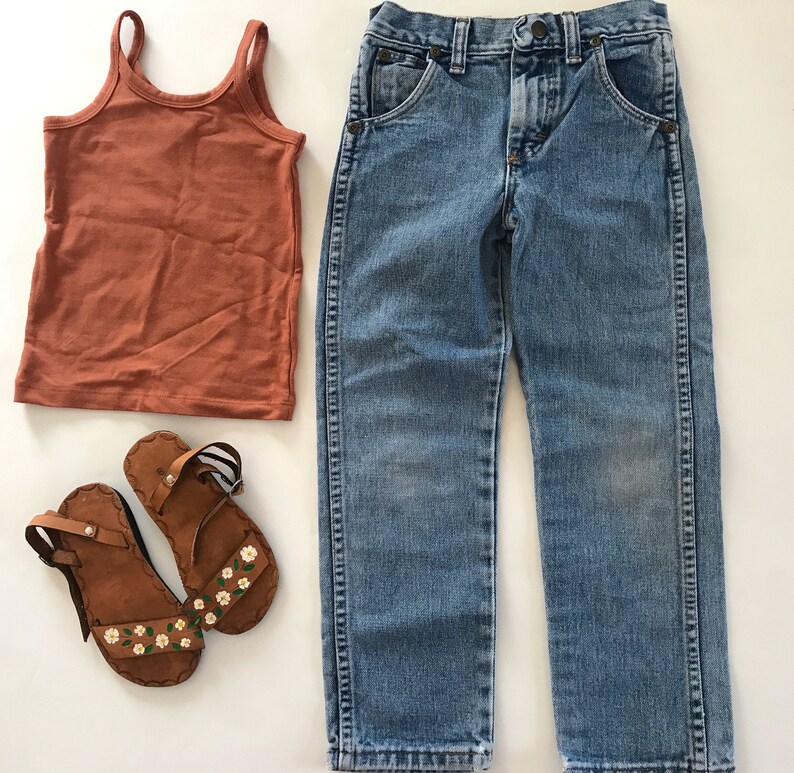May include: A pair of brown leather sandals with white floral designs, a rust-colored tank top, and a pair of light blue denim jeans.