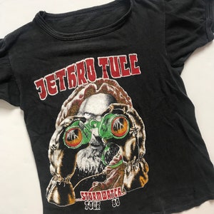 May include: Black vintage t-shirt with a red and white "Jethro Tull" logo and a graphic of a man wearing binoculars. The text "...Stormwatch... Tour '80" is printed below the graphic.