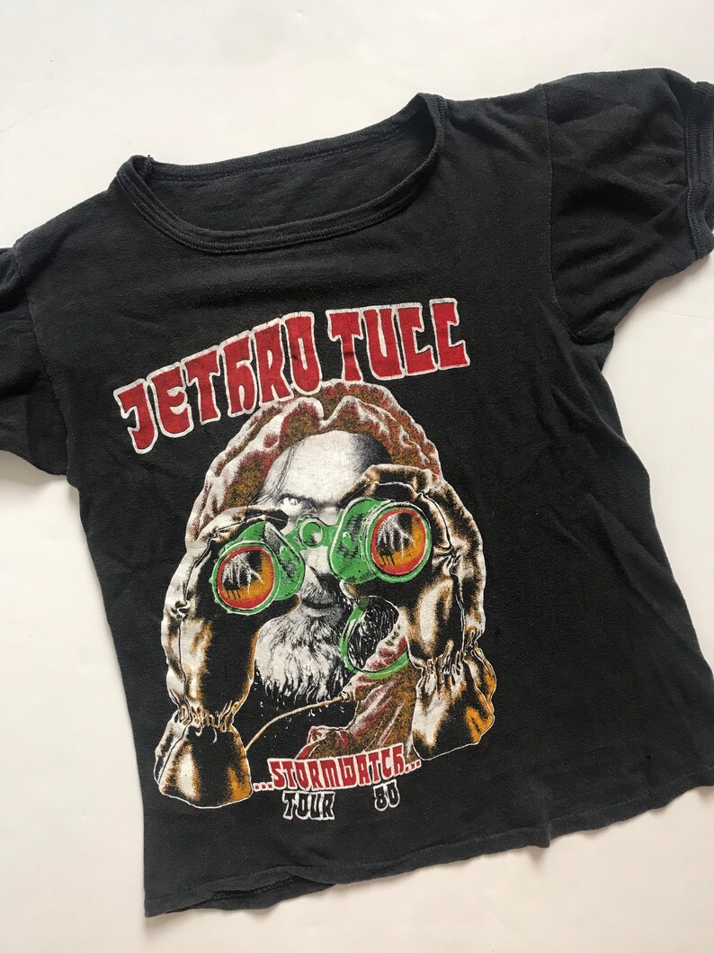 May include: Black vintage t-shirt with a red and white "Jethro Tull" logo and a graphic of a man wearing binoculars. The text "...Stormwatch... Tour '80" is printed below the graphic.