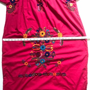 May include: A red embroidered cotton dress with colourful floral designs. The dress has a round neckline and short sleeves. The dress is measured with a tape measure, showing a length of 63.5 cm.
