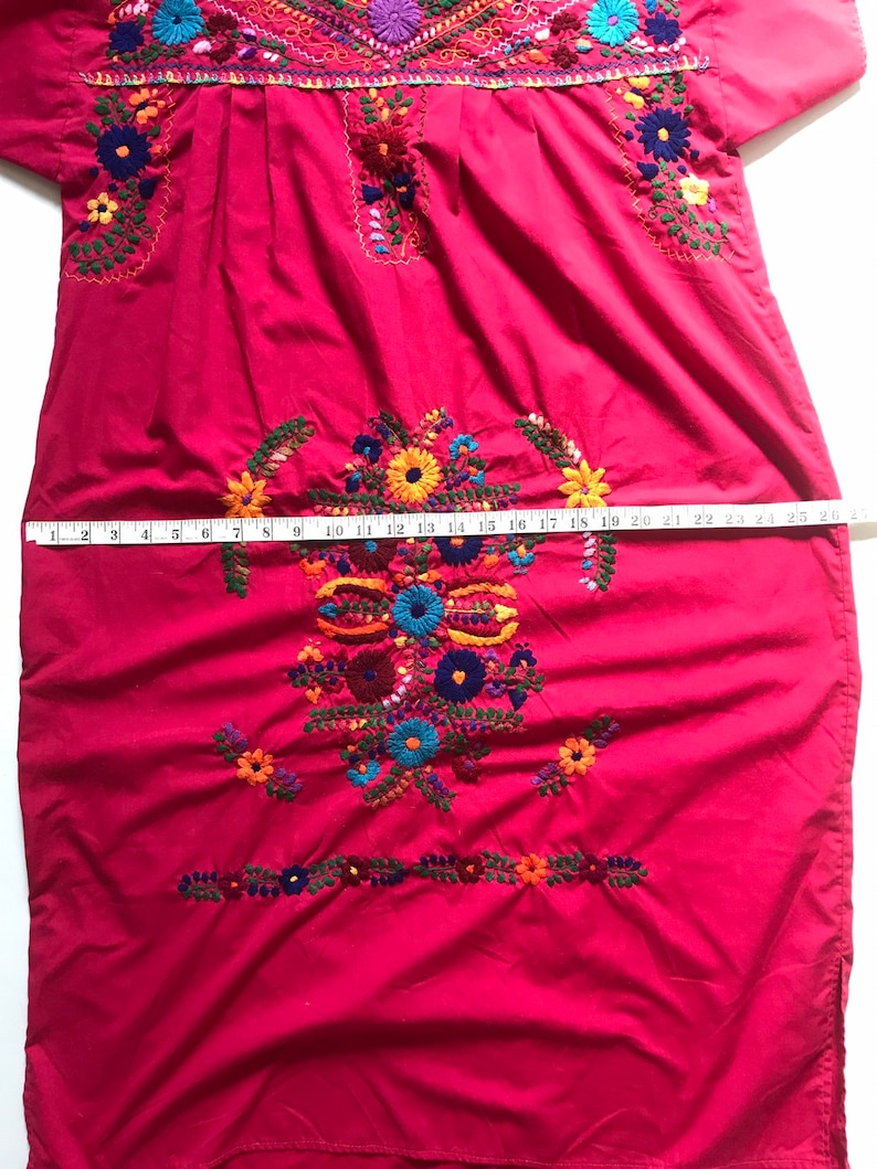 May include: A red embroidered cotton dress with colourful floral designs. The dress has a round neckline and short sleeves. The dress is measured with a tape measure, showing a length of 63.5 cm.