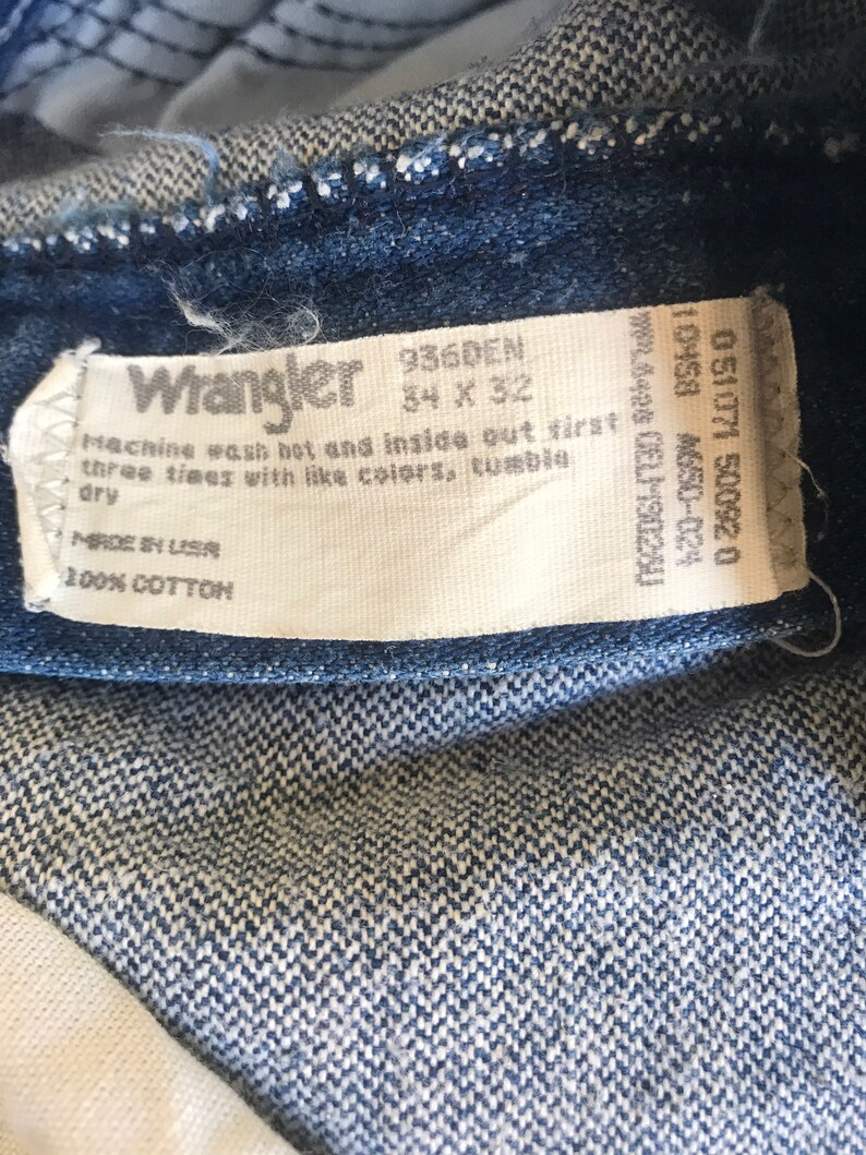 May include: A white tag with black text that reads "Wrangler 936DEN 34 x 32" and "Machine wash hot and inside out first three times with like colors, tumble dry" and "Made in USA 100% Cotton".