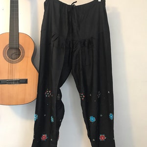 May include: Black pants with embroidered floral details in shades of blue, red, and green. The pants have a drawstring waist and a wide leg.