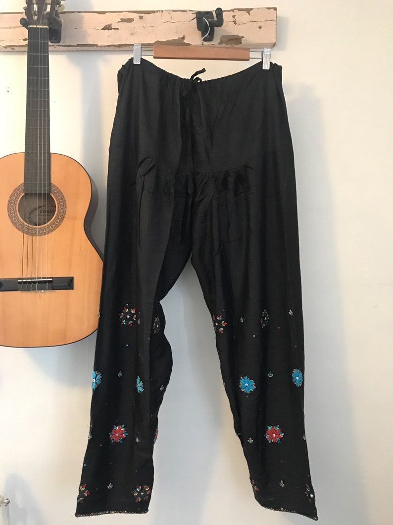 May include: Black pants with embroidered floral details in shades of blue, red, and green. The pants have a drawstring waist and a wide leg.