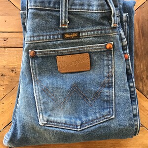 May include: A pair of blue denim jeans folded in half, showing the back pockets. The jeans have a brown leather patch with the word "Wrangler" on it. The jeans also have a small brown leather patch with the word "Wrangler" on it.