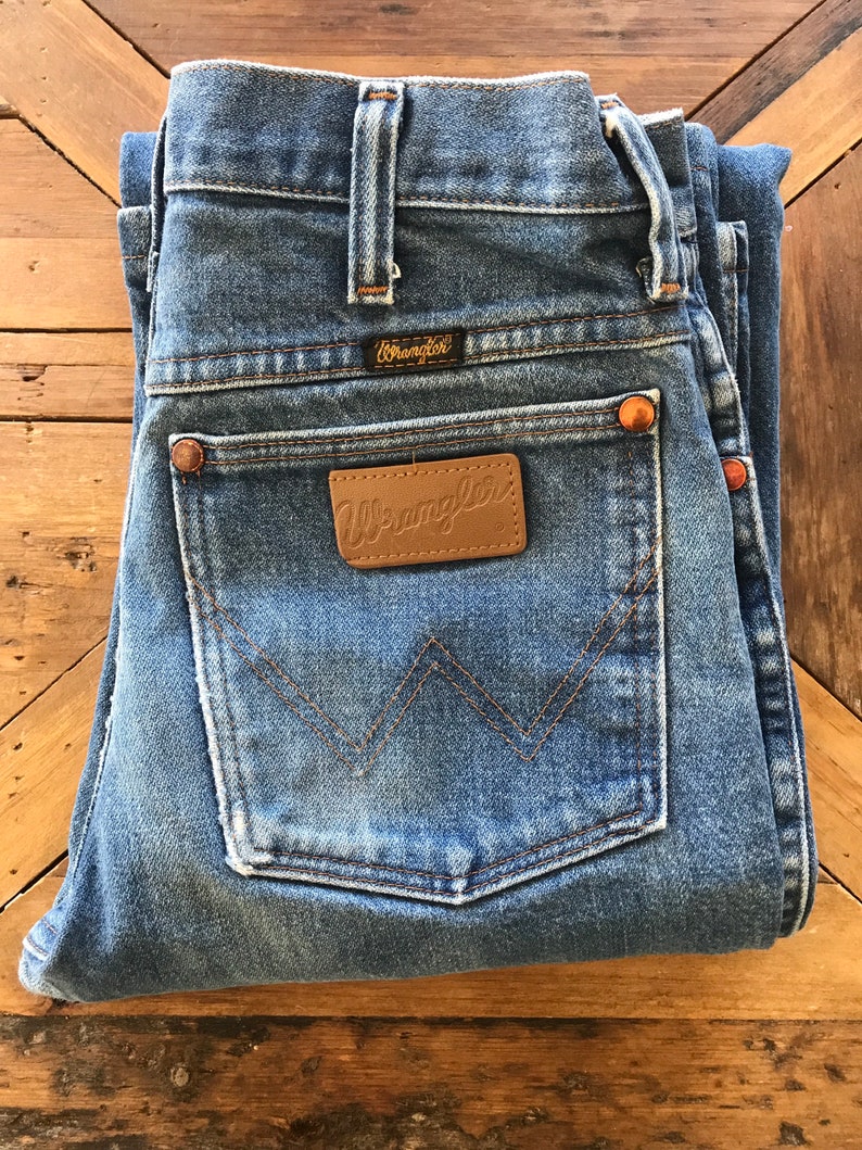 May include: A pair of blue denim jeans folded in half, showing the back pockets. The jeans have a brown leather patch with the word "Wrangler" on it. The jeans also have a small brown leather patch with the word "Wrangler" on it.