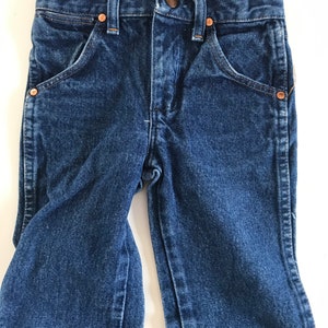 May include: Close-up of a pair of dark blue denim jeans with copper-colored rivets and a button closure. The jeans have classic five-pocket styling and are measured with a ruler. The waist measures approximately 11 inches.