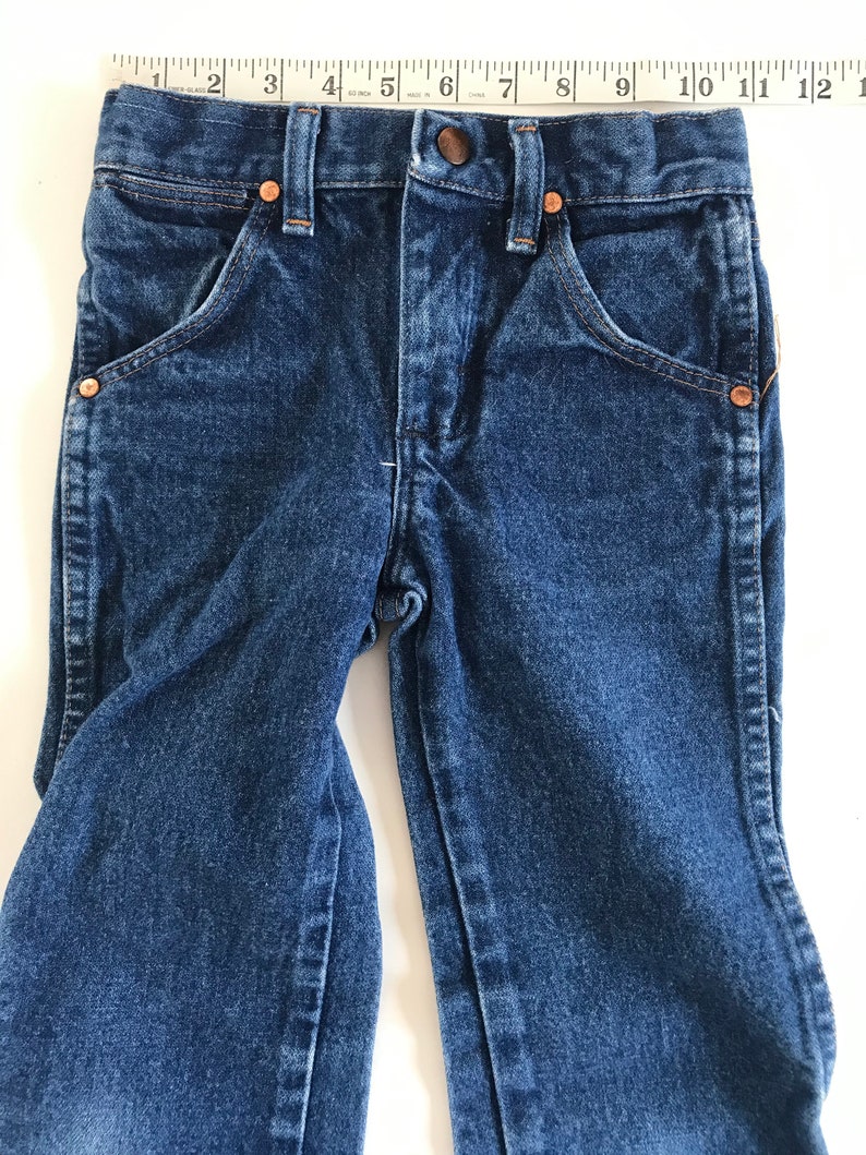 May include: Close-up of a pair of dark blue denim jeans with copper-colored rivets and a button closure. The jeans have classic five-pocket styling and are measured with a ruler. The waist measures approximately 11 inches.