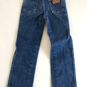 May include: Back view of blue denim jeans with two back pockets and a brown leather patch. The jeans have a Wrangler label and are designed for children. The denim is a medium wash, and the jeans are laid flat.