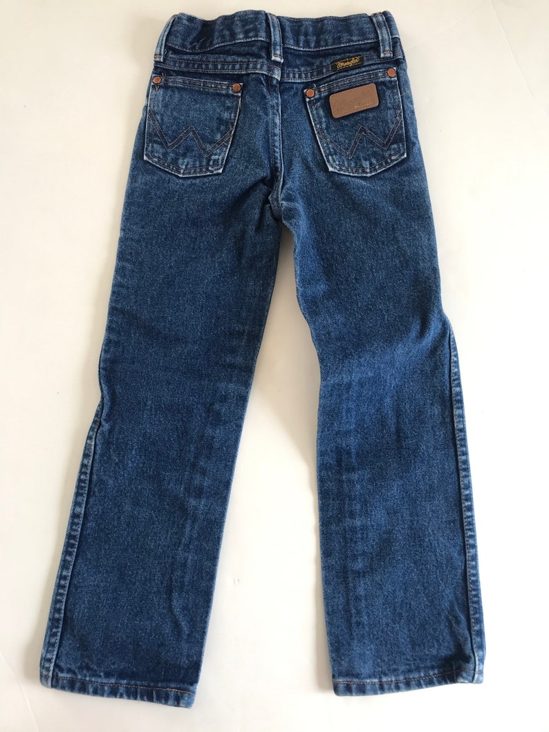 May include: Back view of blue denim jeans with two back pockets and a brown leather patch. The jeans have a Wrangler label and are designed for children. The denim is a medium wash, and the jeans are laid flat.