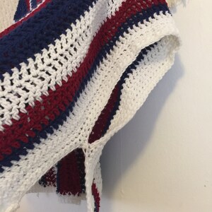 May include: A white, red, and blue striped crochet poncho with a fringe detail. The poncho is made with a loose, open stitch.