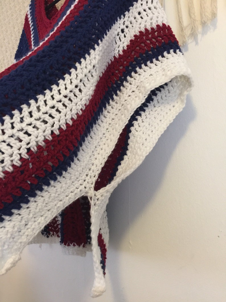 May include: A white, red, and blue striped crochet poncho with a fringe detail. The poncho is made with a loose, open stitch.