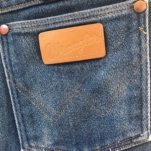 May include: A close-up of a pair of blue denim jeans with a brown leather patch that says "Wrangler" on it. The jeans have a classic five-pocket style with a back pocket that has a distinctive "W" shape.