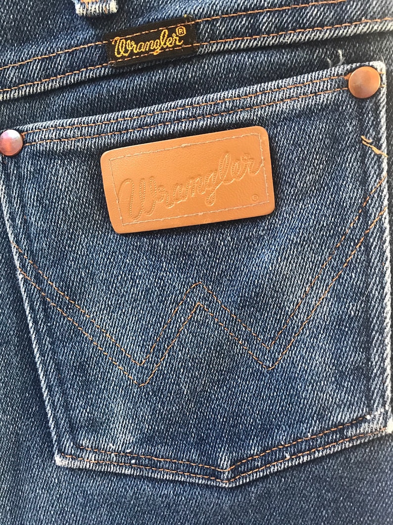 May include: A close-up of a pair of blue denim jeans with a brown leather patch that says "Wrangler" on it. The jeans have a classic five-pocket style with a back pocket that has a distinctive "W" shape.