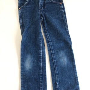 May include: Dark blue denim jeans with classic five-pocket styling. Features include copper rivets, a button closure, and a straight leg. The jeans are designed for children and are displayed on a white surface.