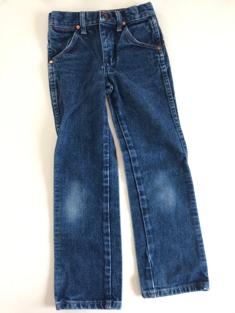 May include: Dark blue denim jeans with classic five-pocket styling. Features include copper rivets, a button closure, and a straight leg. The jeans are designed for children and are displayed on a white surface.