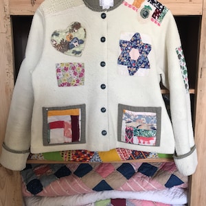 May include: Cream-colored, button-down jacket with patchwork details. Features a heart-shaped patch with a woodland scene, a six-pointed star, and floral and geometric fabric squares. The jacket is hanging on a wooden hanger.