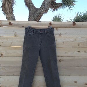 May include: A pair of dark brown straight-leg jeans hanging on a wooden hanger against a wood panel background.