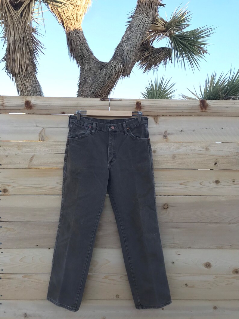 May include: A pair of dark brown straight-leg jeans hanging on a wooden hanger against a wood panel background.
