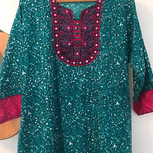 Puede incluir: T&uacute;nica azul turquesa con un estampado floral rosa y blanco y un escote decorativo con adornos de espejo. El escote tiene un ribete rosa y una peque&ntilde;a etiqueta que dice "Ethnic House".
