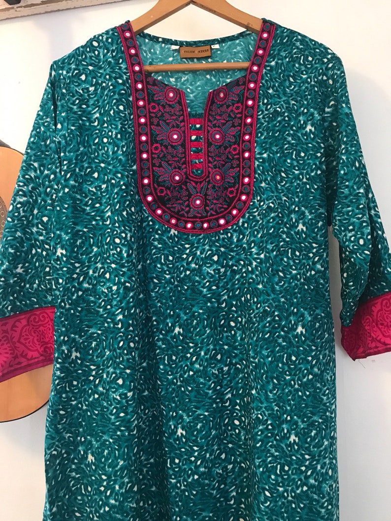 Puede incluir: T&uacute;nica azul turquesa con un estampado floral rosa y blanco y un escote decorativo con adornos de espejo. El escote tiene un ribete rosa y una peque&ntilde;a etiqueta que dice "Ethnic House".