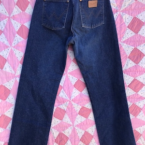 Może przedstawiać: Dżinsy z ciemnego denimu z brązową sk&oacute;rzaną naszywką na tylnej kieszeni z napisem "Wrangler".