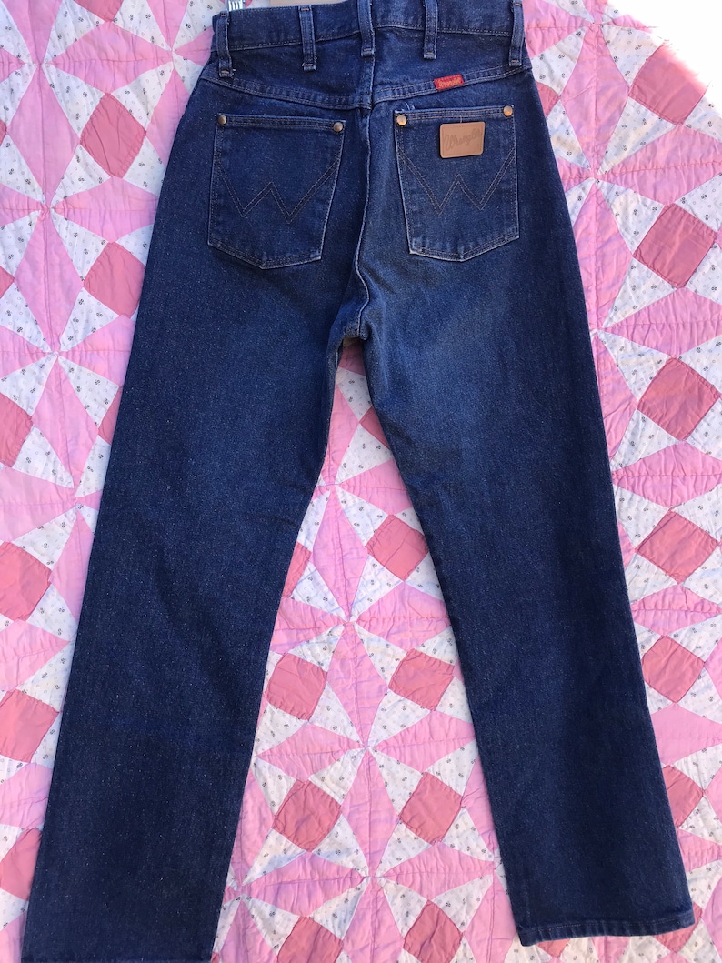 Może przedstawiać: Dżinsy z ciemnego denimu z brązową sk&oacute;rzaną naszywką na tylnej kieszeni z napisem "Wrangler".